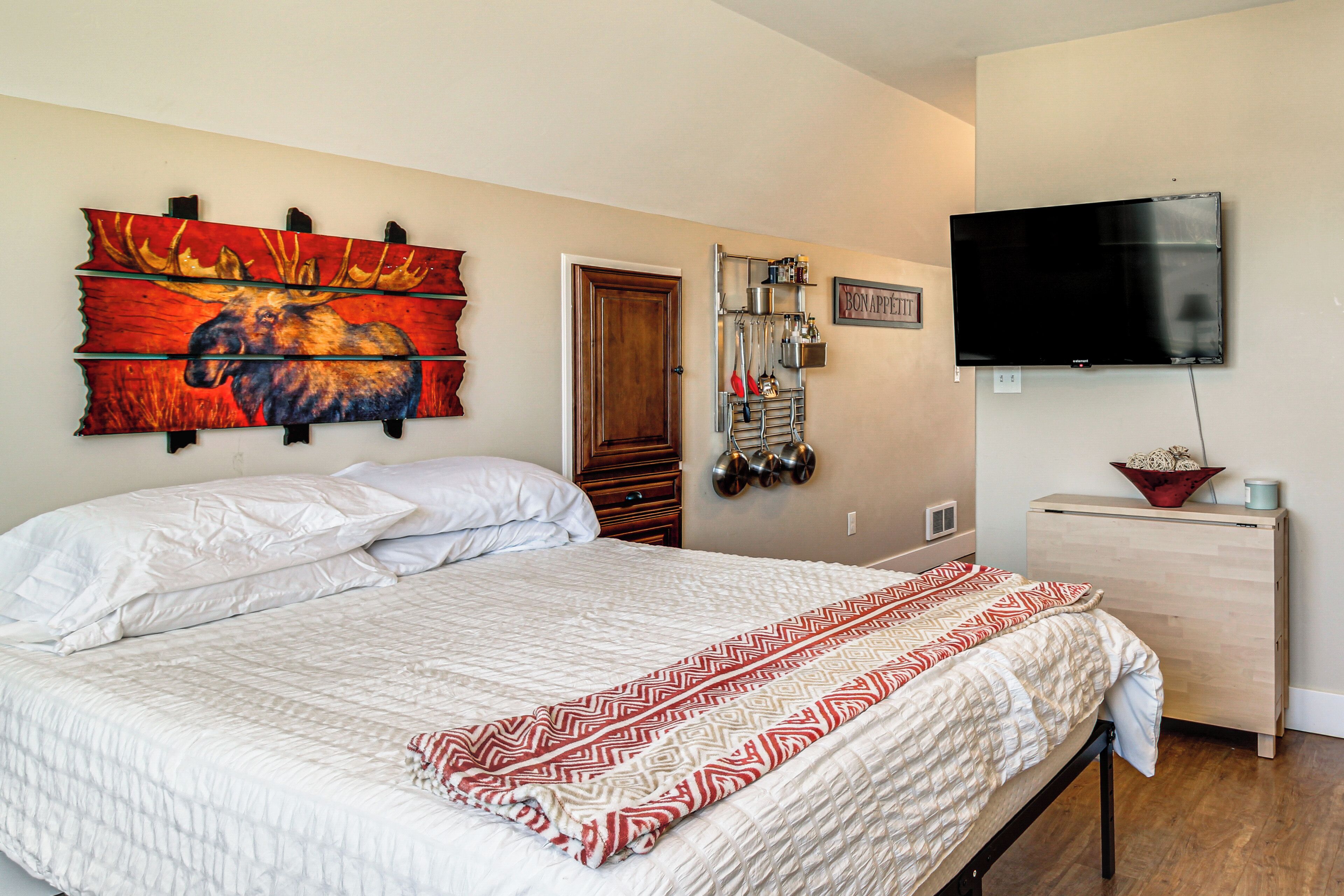 A bedroom with a bed, a mounted TV, a wall-mounted shelf with kitchen utensils, and a door.