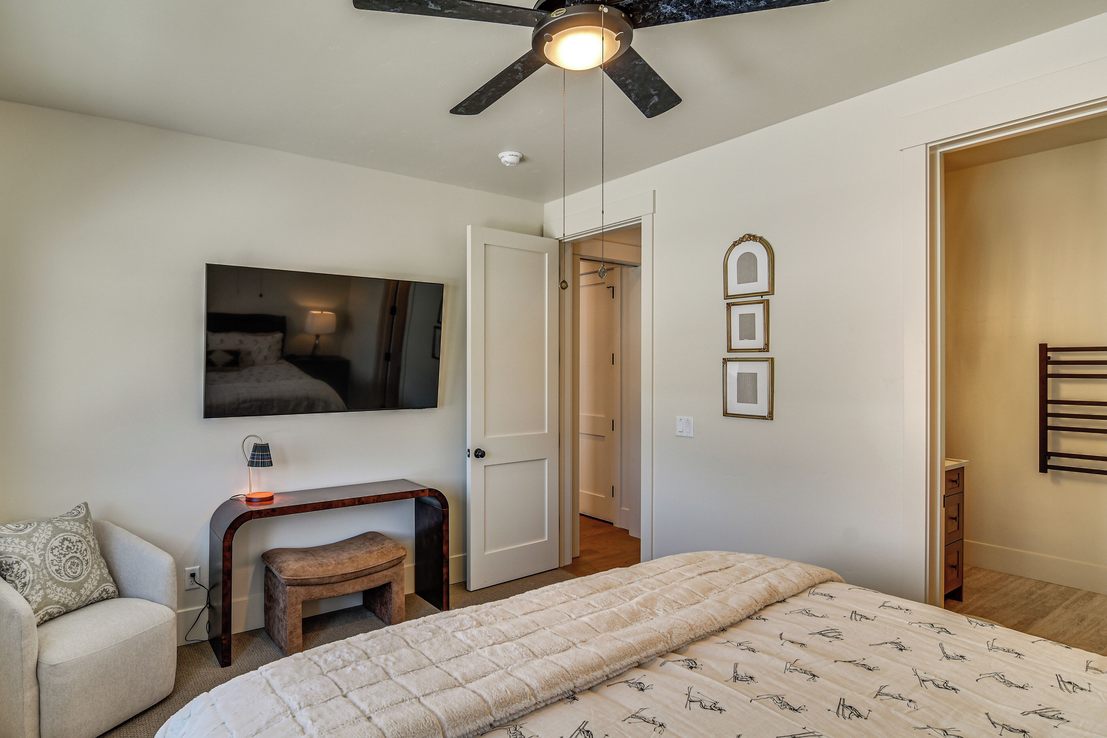 Une chambre à coucher comprenant un lit, un bureau, une chaise, un ventilateur de plafond, une télévision et une porte donnant sur une autre pièce.