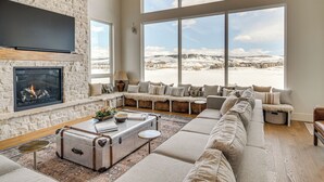 Un salón amplio con pared de piedra, chimenea, un ventanal con vistas nevadas, una zona de estar acogedora y una maleta antigua.