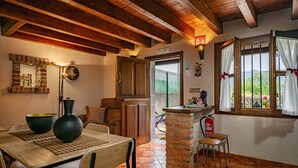 A rustic interior with a wooden table, chairs, a brick column, and a view through a window.