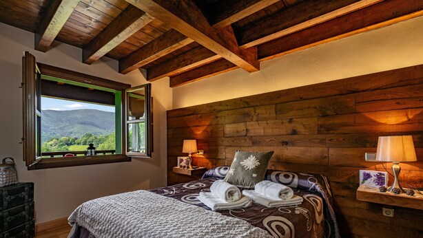 A bedroom with a wooden ceiling, a large bed with a patterned bedspread, two bedside lamps, and a window with a view of mountains.