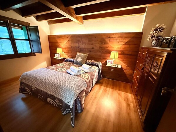 Un dormitorio con cabecera de madera, dos mesitas de noche con lámparas, una cama con ropa de cama estampada y un aparador con objetos decorativos.