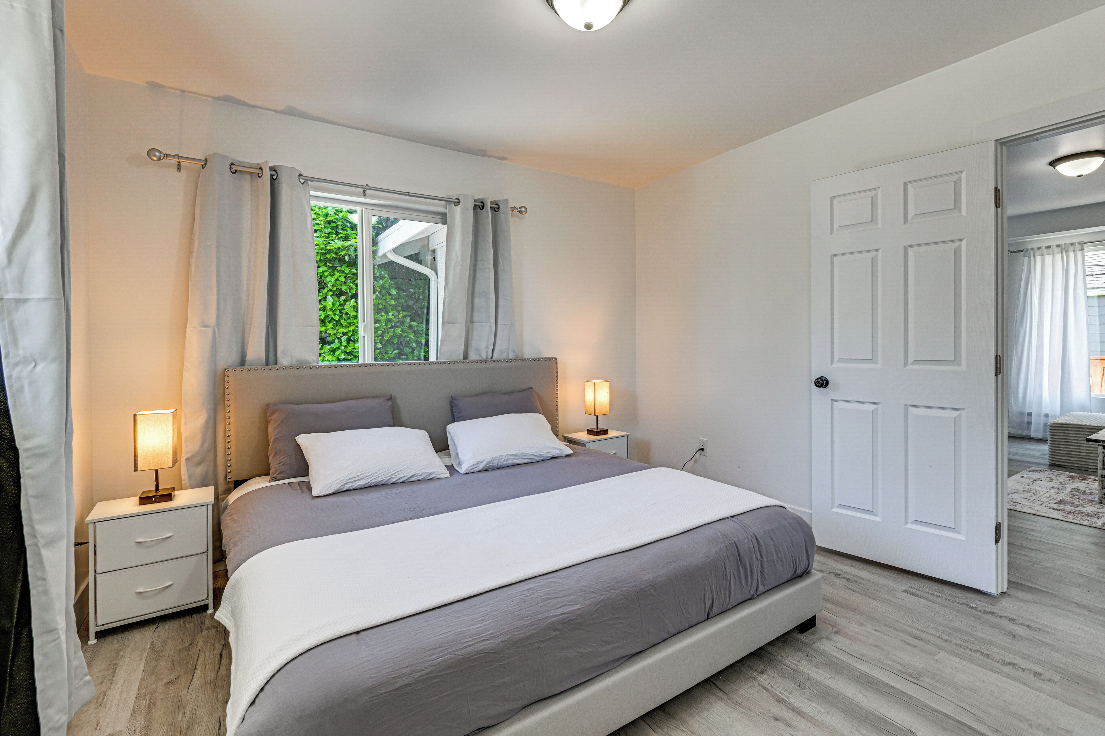 A bedroom with a bed, nightstands, a window with curtains, a door, and a view of greenery outside.