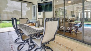 Un patio con mesa y sillas, un televisor montado en la pared y vista al jardín trasero a través de puertas de vidrio.