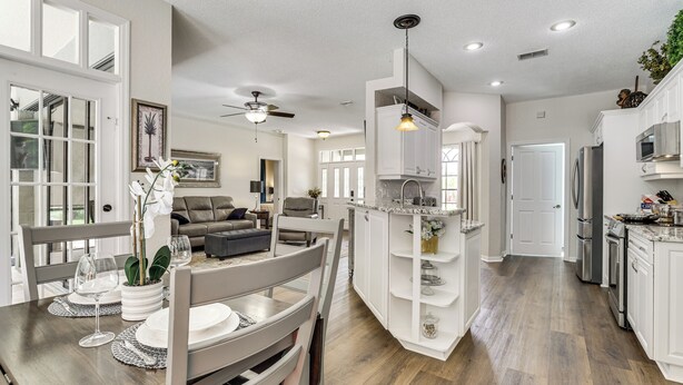 Una cocina amplia con armarios blancos, un comedor con mesa para dos invitados y una sala de estar con sofá y ventilador de techo.