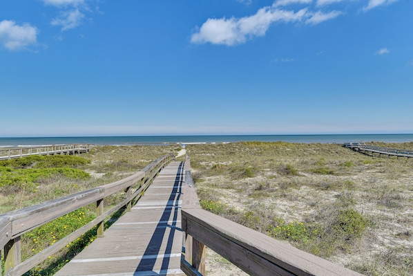 Ein hölzerner Steg führt zu einem Strand mit Dünen und blauem Himmel.