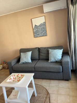 Un canapé gris avec deux coussins, une table blanche avec un livre, et une œuvre d’art abstraite accrochée au mur.