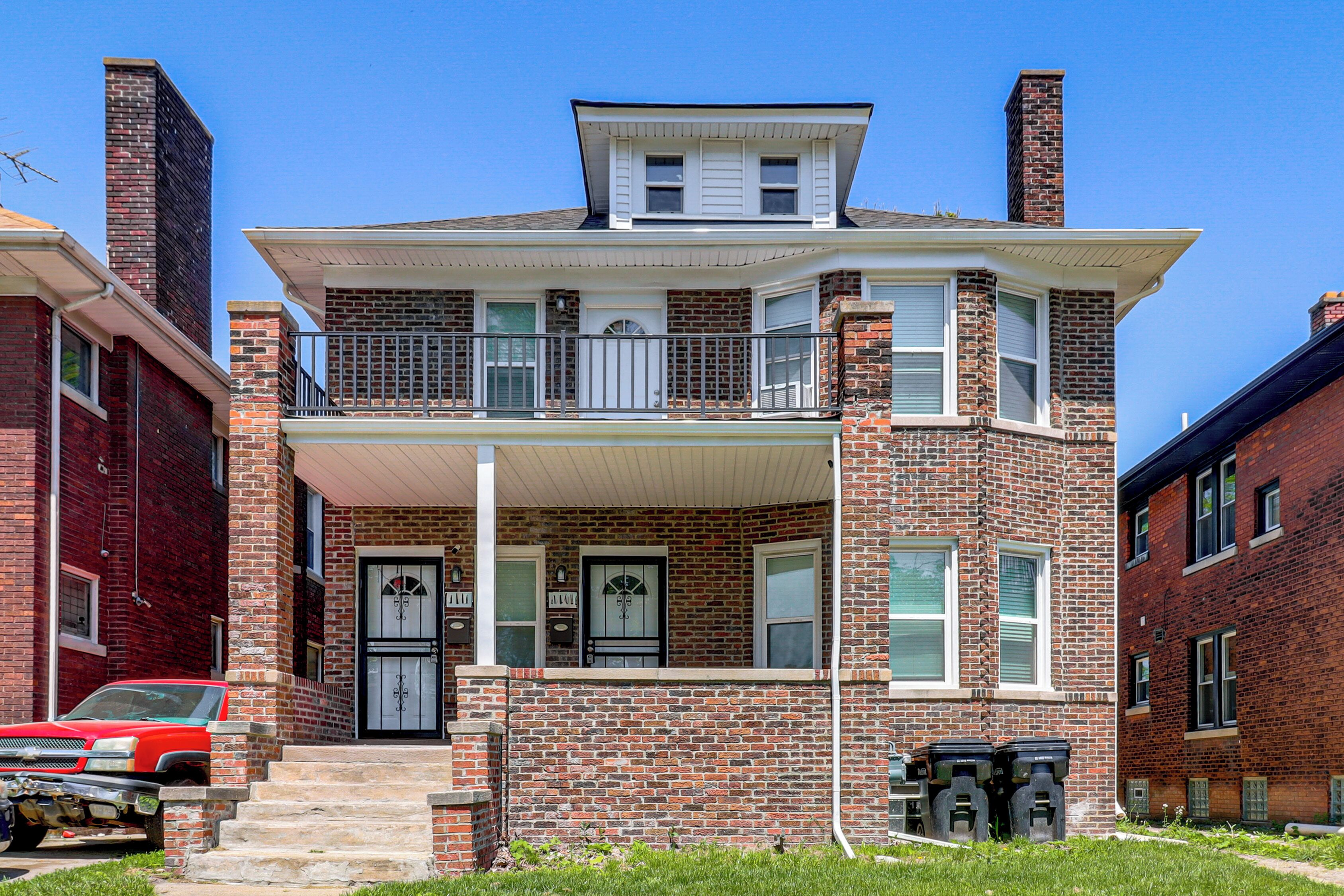 A two-story brick house with a balcony, a front porch, and a garage with a red truck parked outside.
