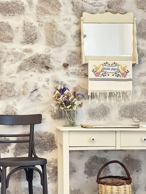Un mur de pierres apparentes, une chaise en bois, un bureau blanc avec des tiroirs, un panier en osier et un miroir agrémenté d’un tissu décoratif.