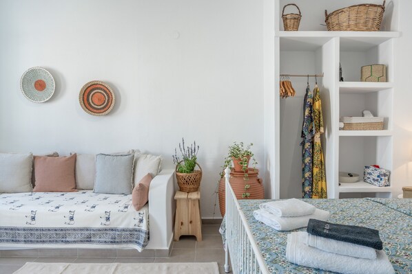 Un dormitorio con una cama, un taburete pequeño, una mesita de noche, un estante con cestas y una planta en maceta.