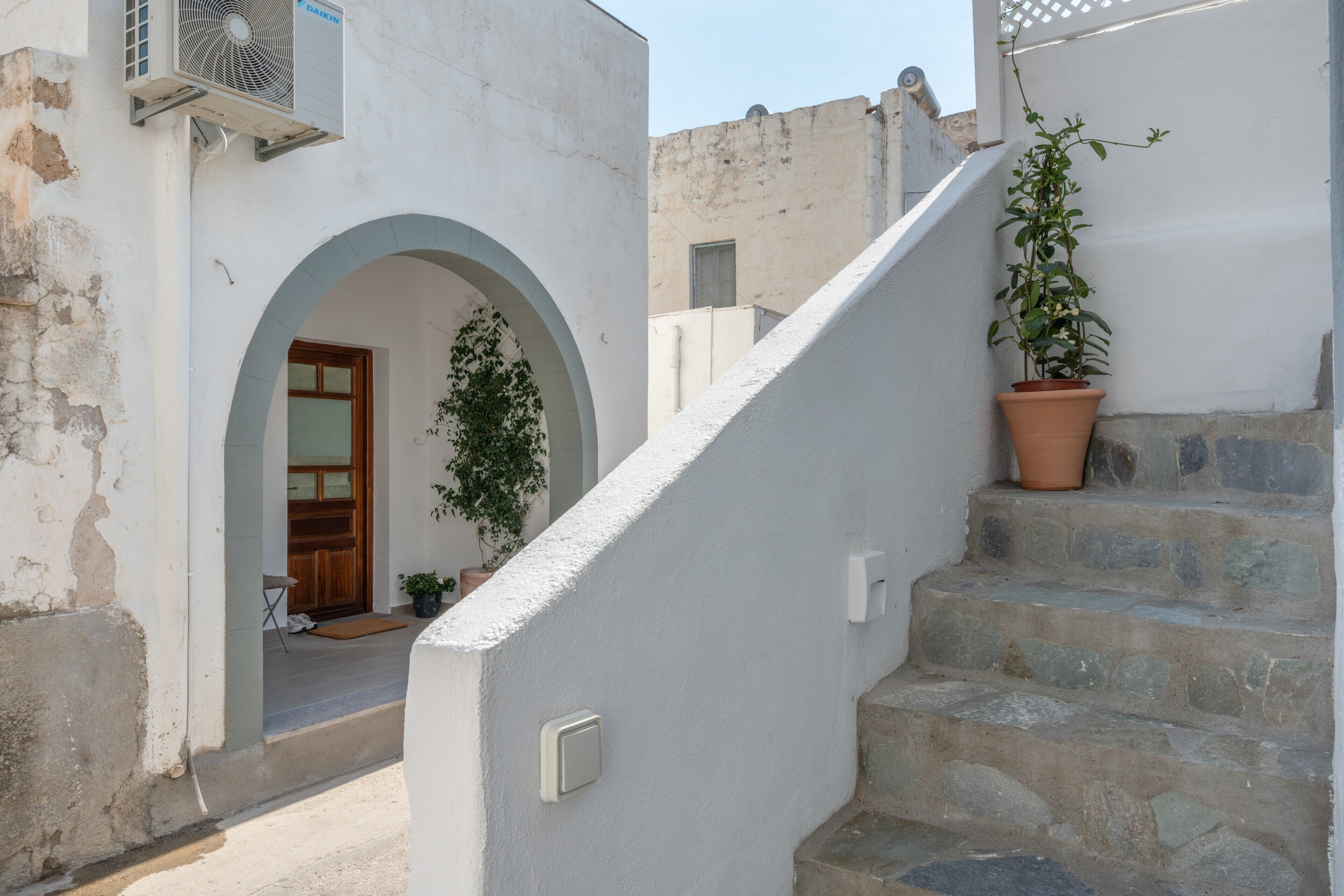 A narrow street with a white building, a wooden door, a potted plant, and a stone staircase.
