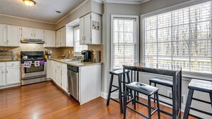 A kitchen with white cabinets, stainless steel appliances, a window with blinds, and a bar stool. - Centrally Located Fayetteville Home: Deck + Yard (Fayetteville)