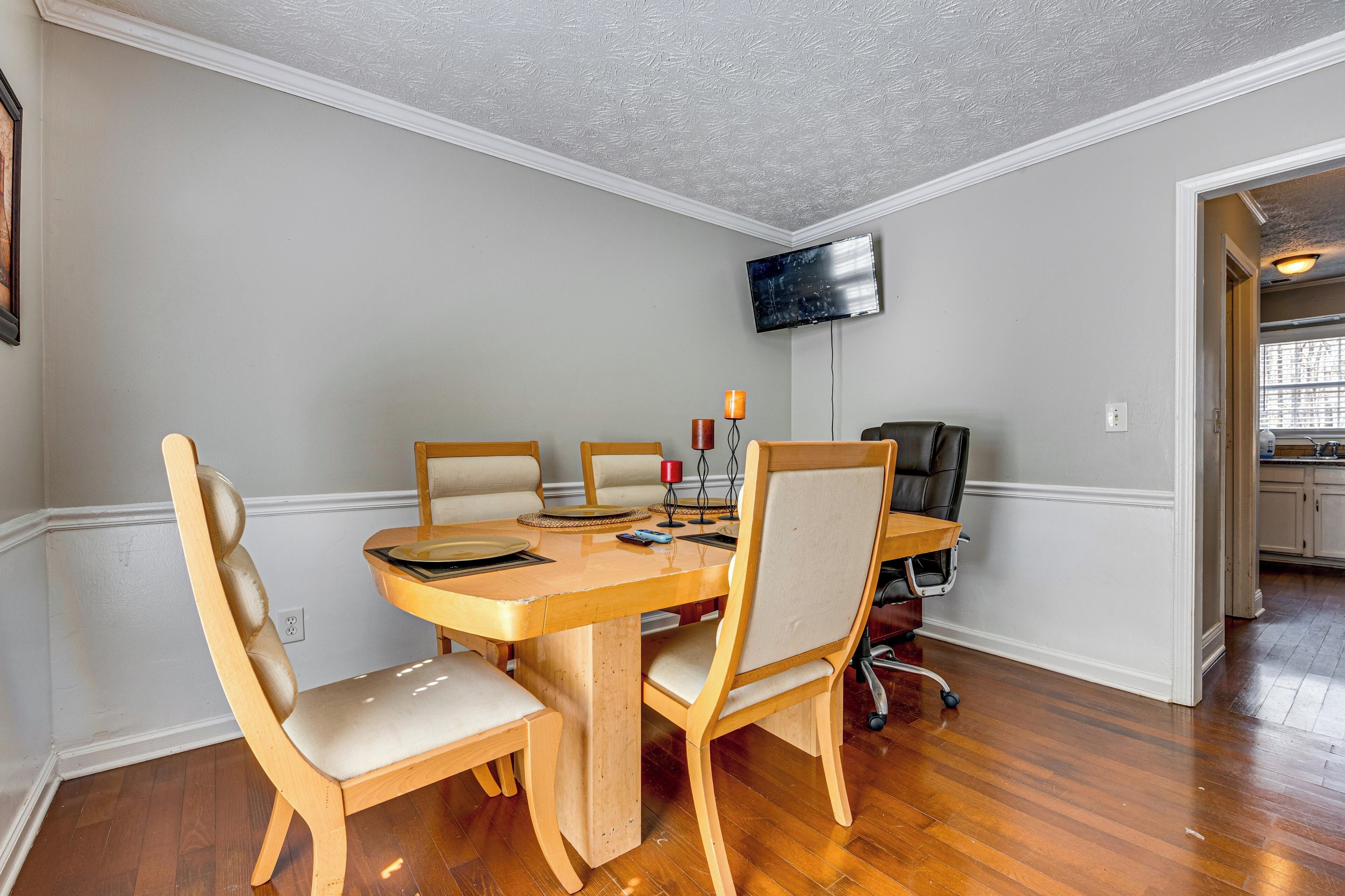 A dining area with a wooden table, four chairs, a television mounted on the wall, and a kitchen visible in the background.