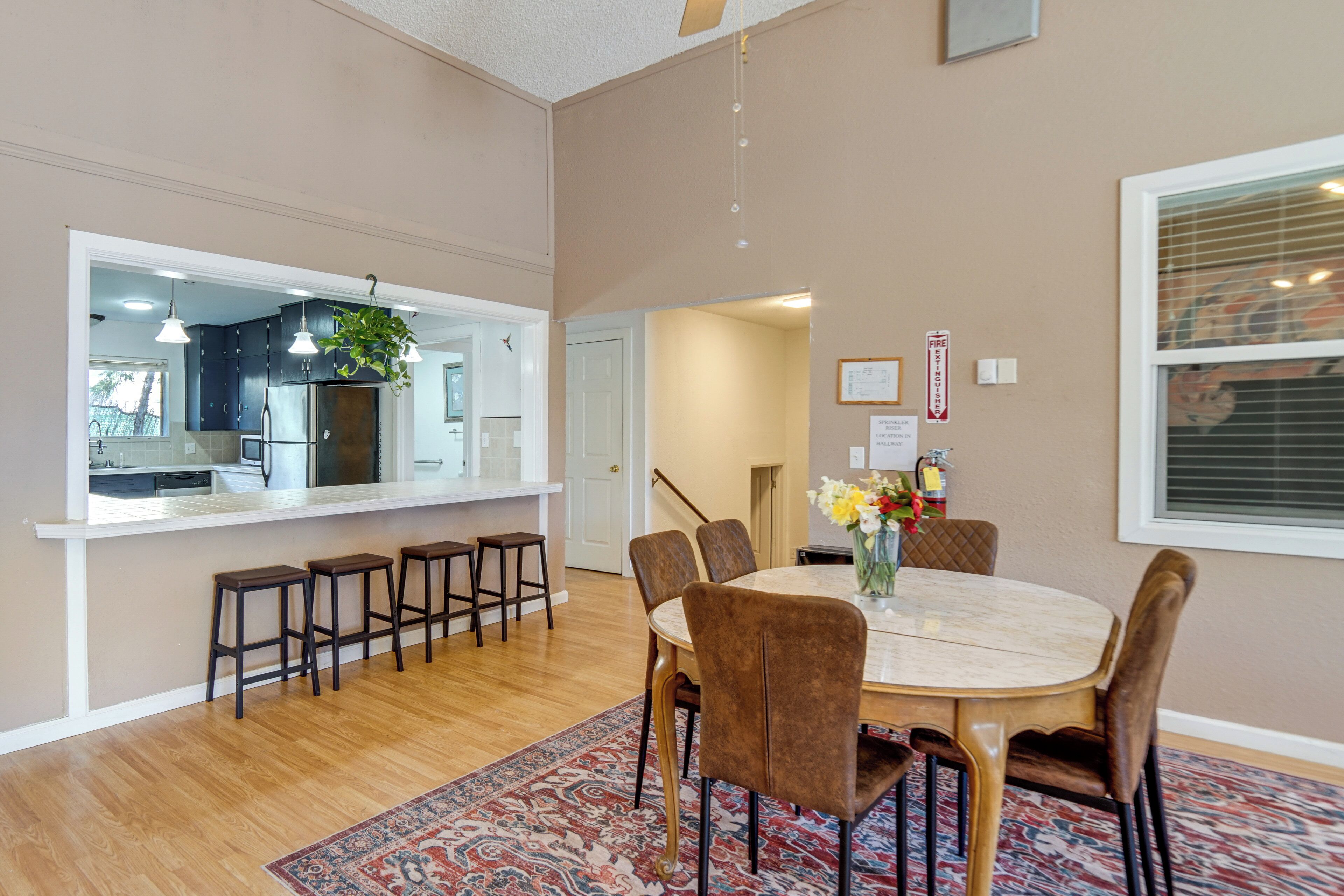 Un coin repas avec une table ronde, six chaises, un tapis à motifs et une cuisine visible à travers une porte ouverte.