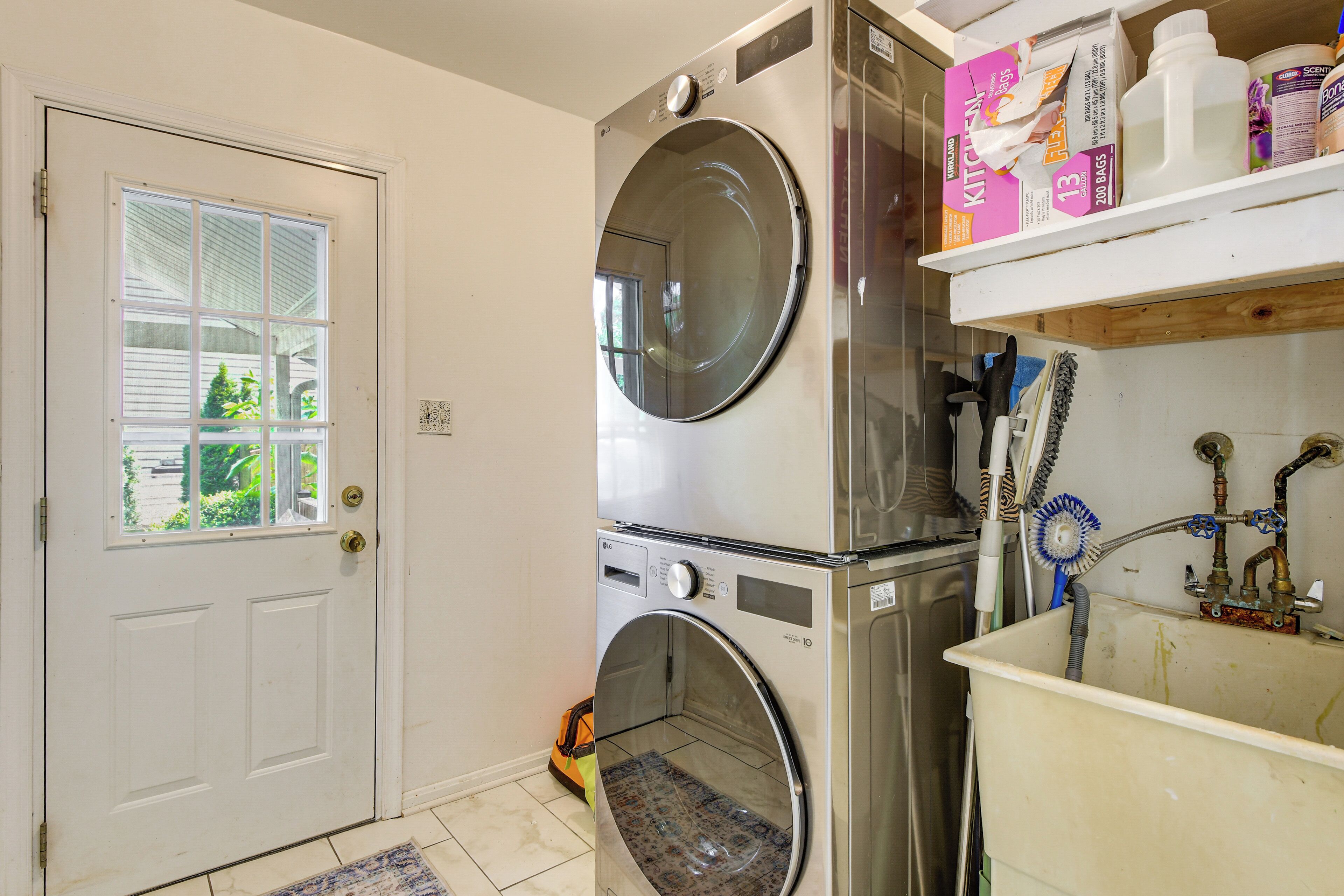 Un espace buanderie avec des machines à laver et des sèche-linge superposés, une porte avec une fenêtre et une étagère contenant des produits de nettoyage.