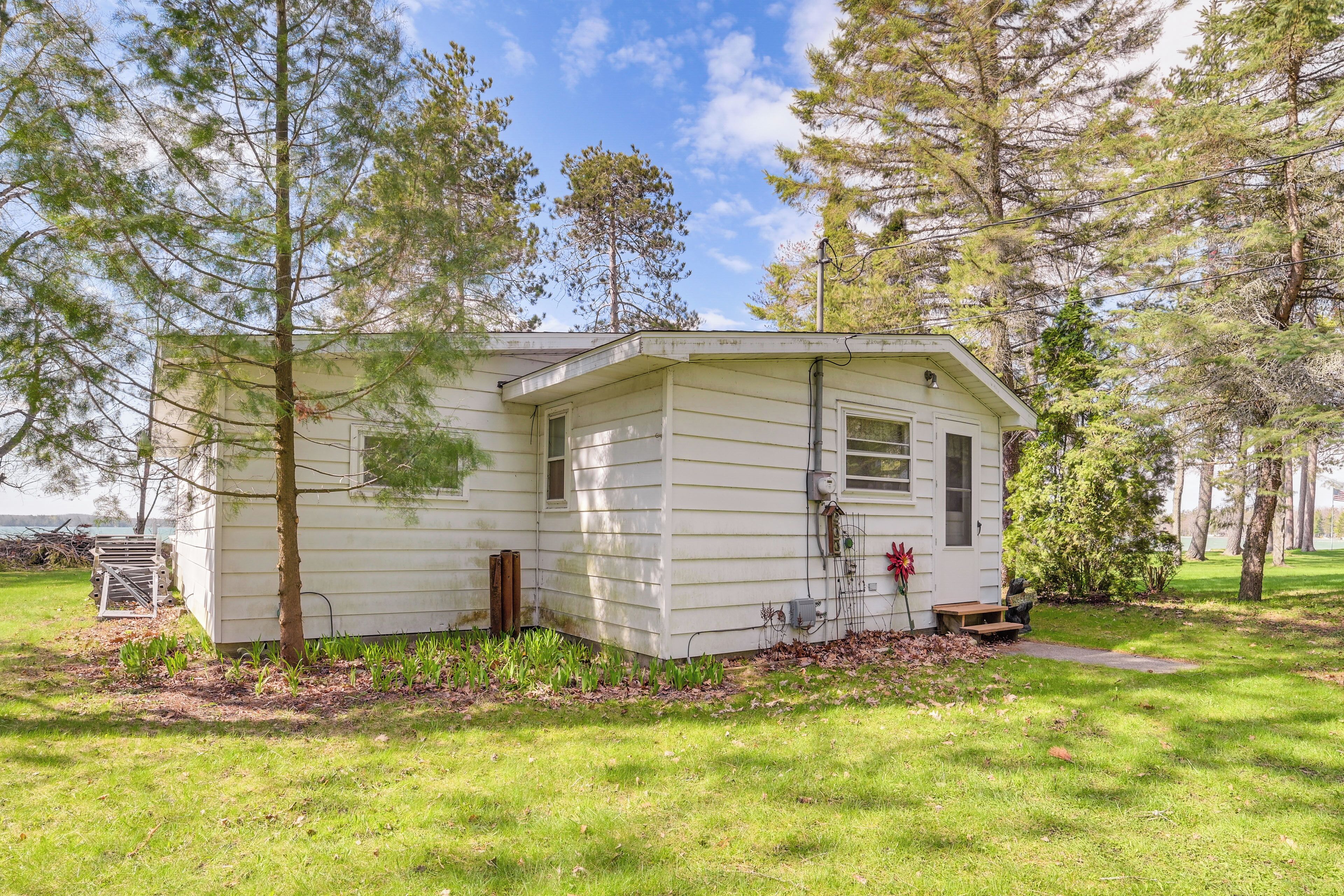 A small, single-story house with a sloped roof, a front porch, and a small garden area with grass and a few trees.