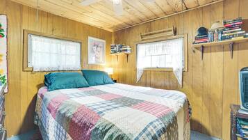 A wooden-paneled room with a bed, two windows, a ceiling fan, and a shelf with books.