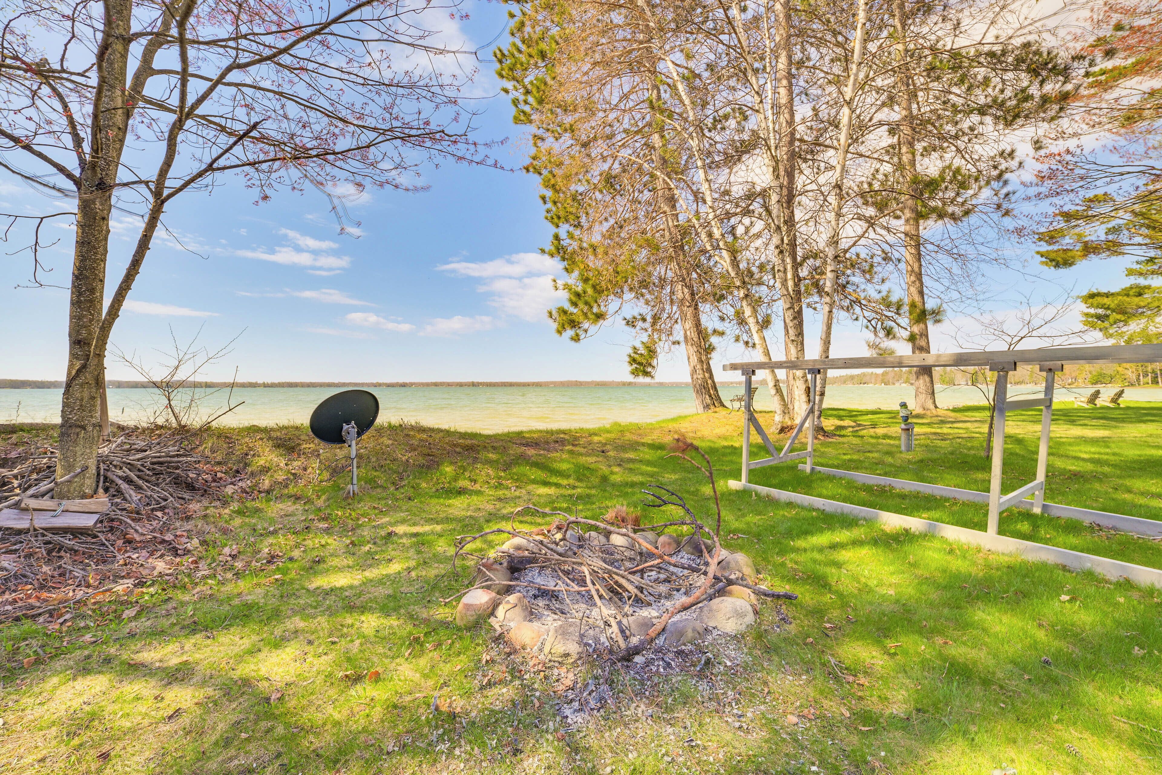 A grassy area with a fire pit, trees, and a bench by a body of water.