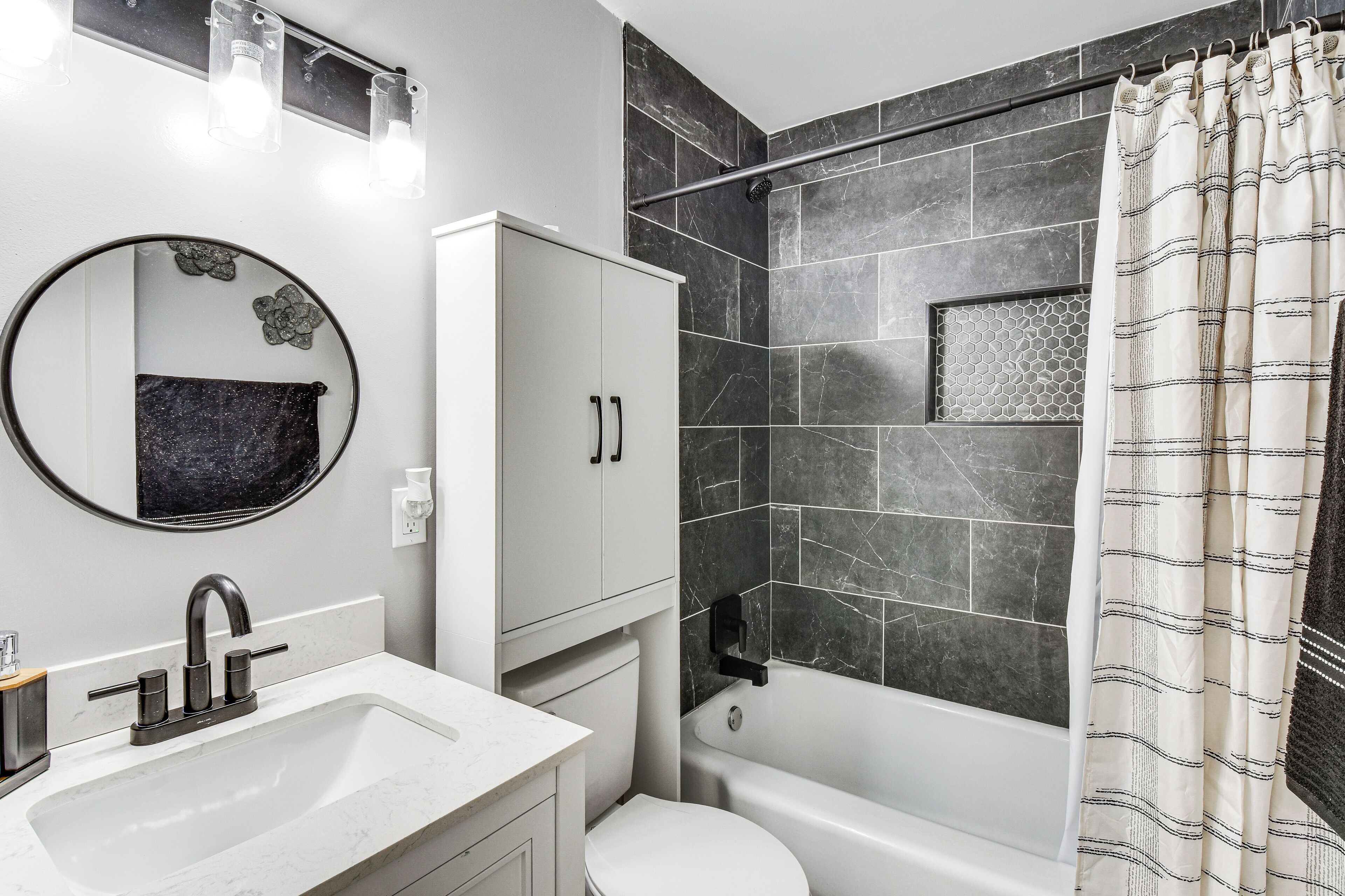 A modern bathroom with a white sink, a toilet, a bathtub, a shower with dark tiles, and a mirror with a flower decoration.