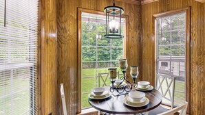 A wooden-paneled room with a round table set for dining, a chandelier, and a view of a green landscape through large windows. - Walkable Location! Peaceful Toccoa Escape w/ Yard (Toccoa)