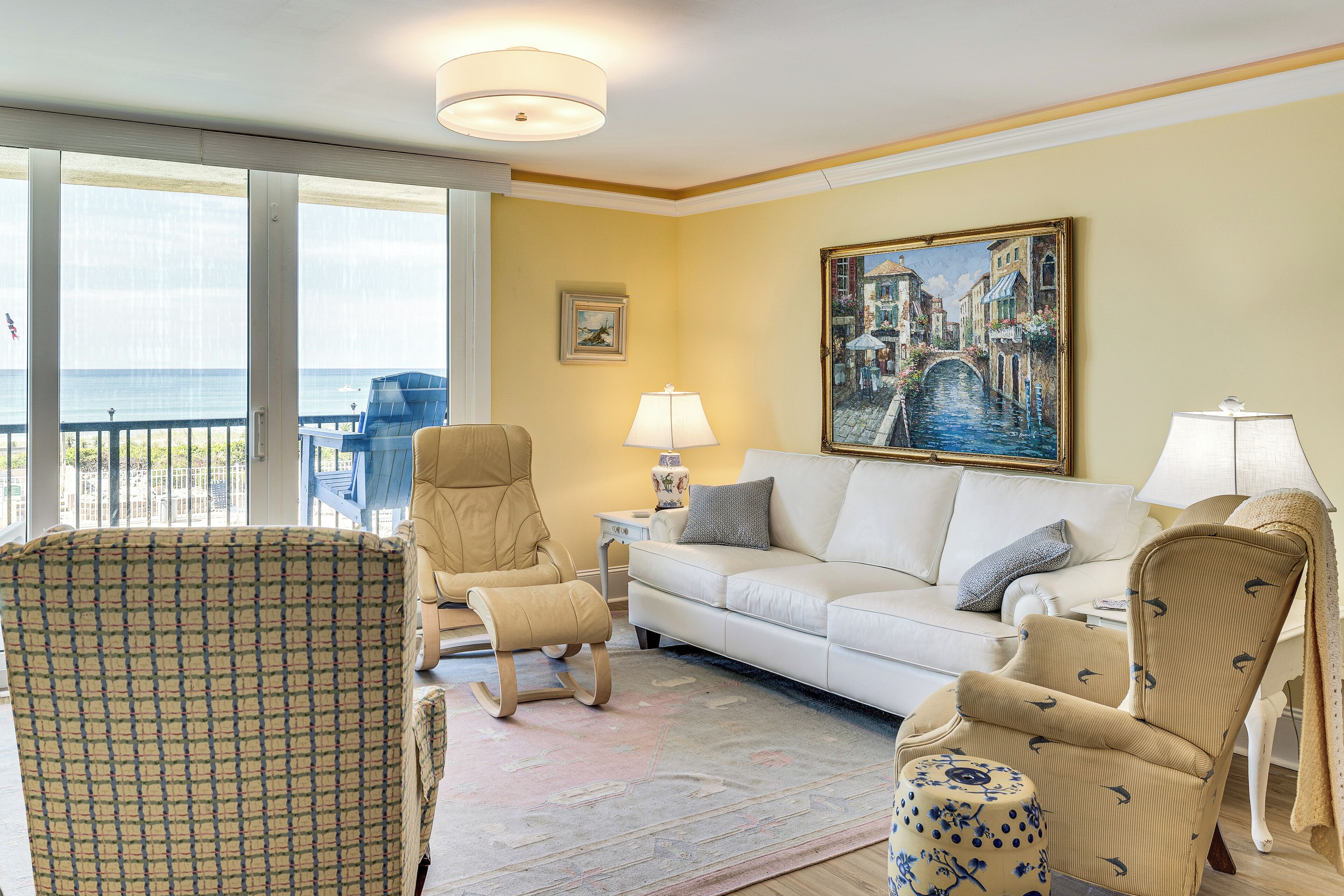 A living room with a sofa, chairs, a patterned armchair, a painting, and a view of the sea.
