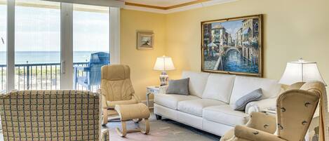 A living room with a sofa, chairs, a patterned armchair, a painting, and a view of the sea.