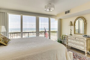 Quarto com uma cama grande, cômoda, espelho e vista para o oceano através de grandes janelas.