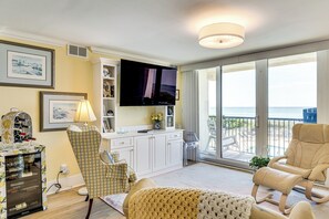 Uma sala de estar com televisão, sofá, poltrona, mesinha e vista para o oceano por grandes janelas.