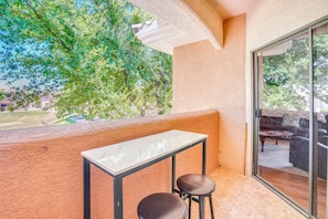 Ein Balkon mit Tisch und zwei Stühlen, eine Glastür zu einer Außenbereich-Sitzgelegenheit und Blick auf Bäume und Gebäude.