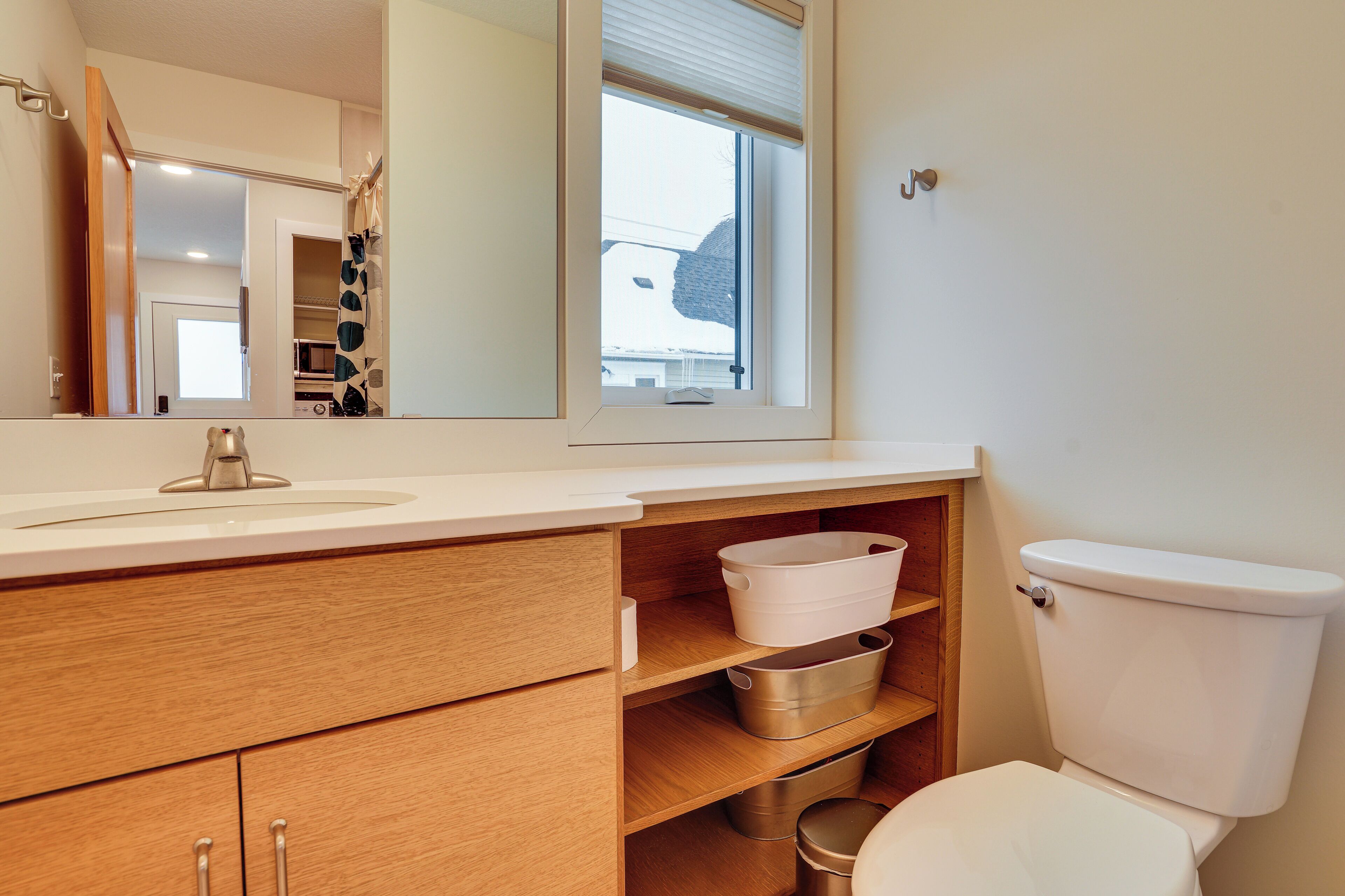 A modern bathroom with a wooden vanity, a white sink, a toilet, and a window with blinds.