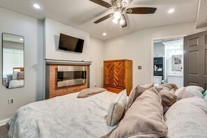 A bedroom with a bed, a television above it, a ceiling fan, a fireplace, and a wardrobe.