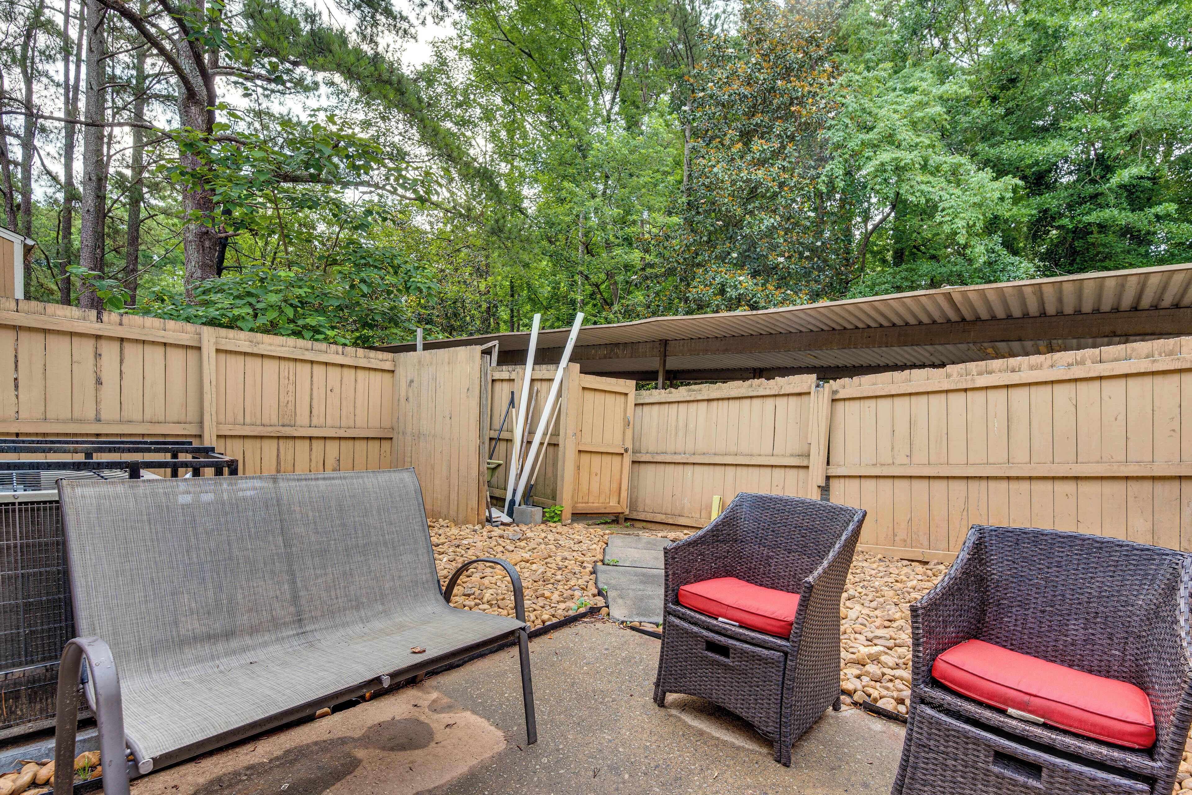 A backyard with a covered patio area, two wicker chairs with red cushions, and a metal bench.