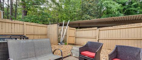 A backyard with a covered patio area, two wicker chairs with red cushions, and a metal bench.