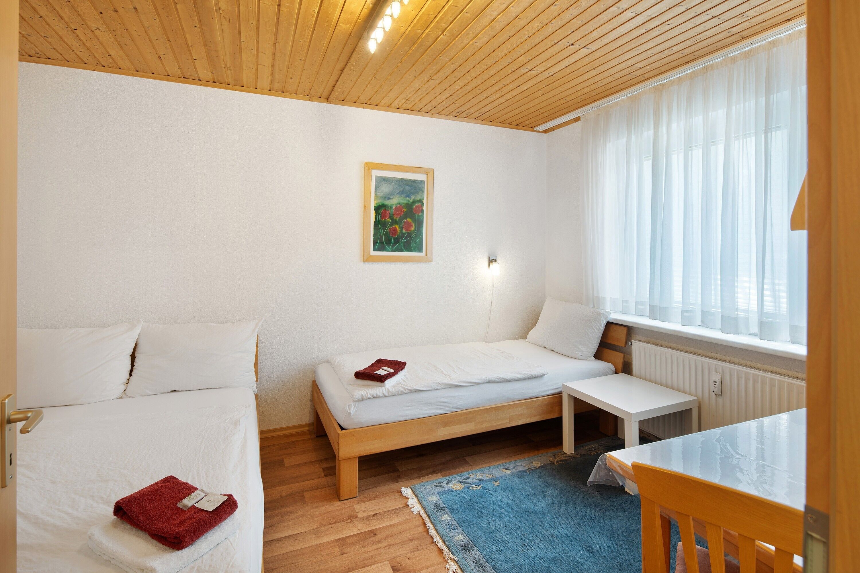 Une chambre avec deux lits, une petite table, une fenêtre avec des rideaux et un tableau au mur.