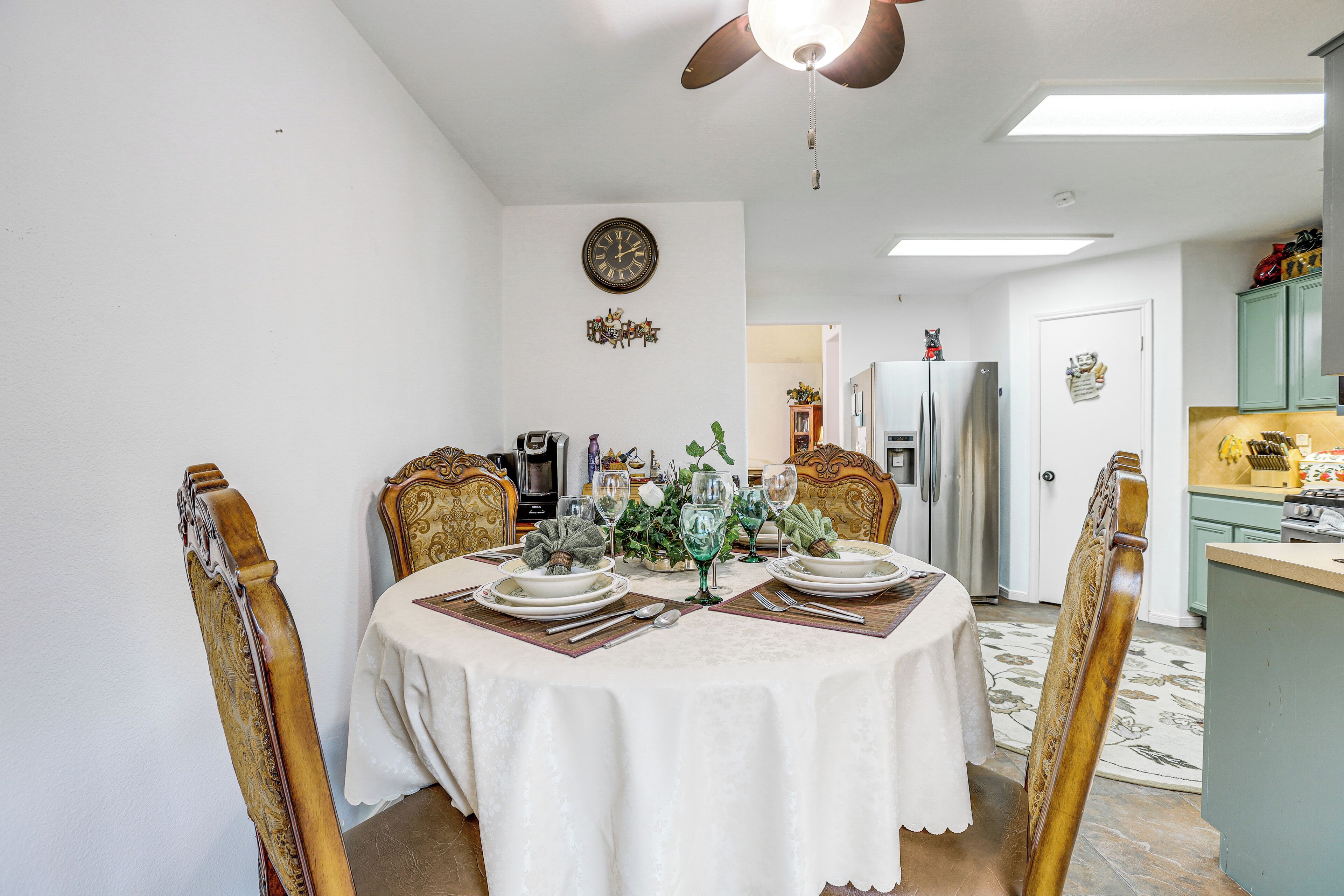 A dining room with a round table set for four, chairs with ornate backs, a ceiling fan, a clock, and a kitchen area with cabinets and appliances.
