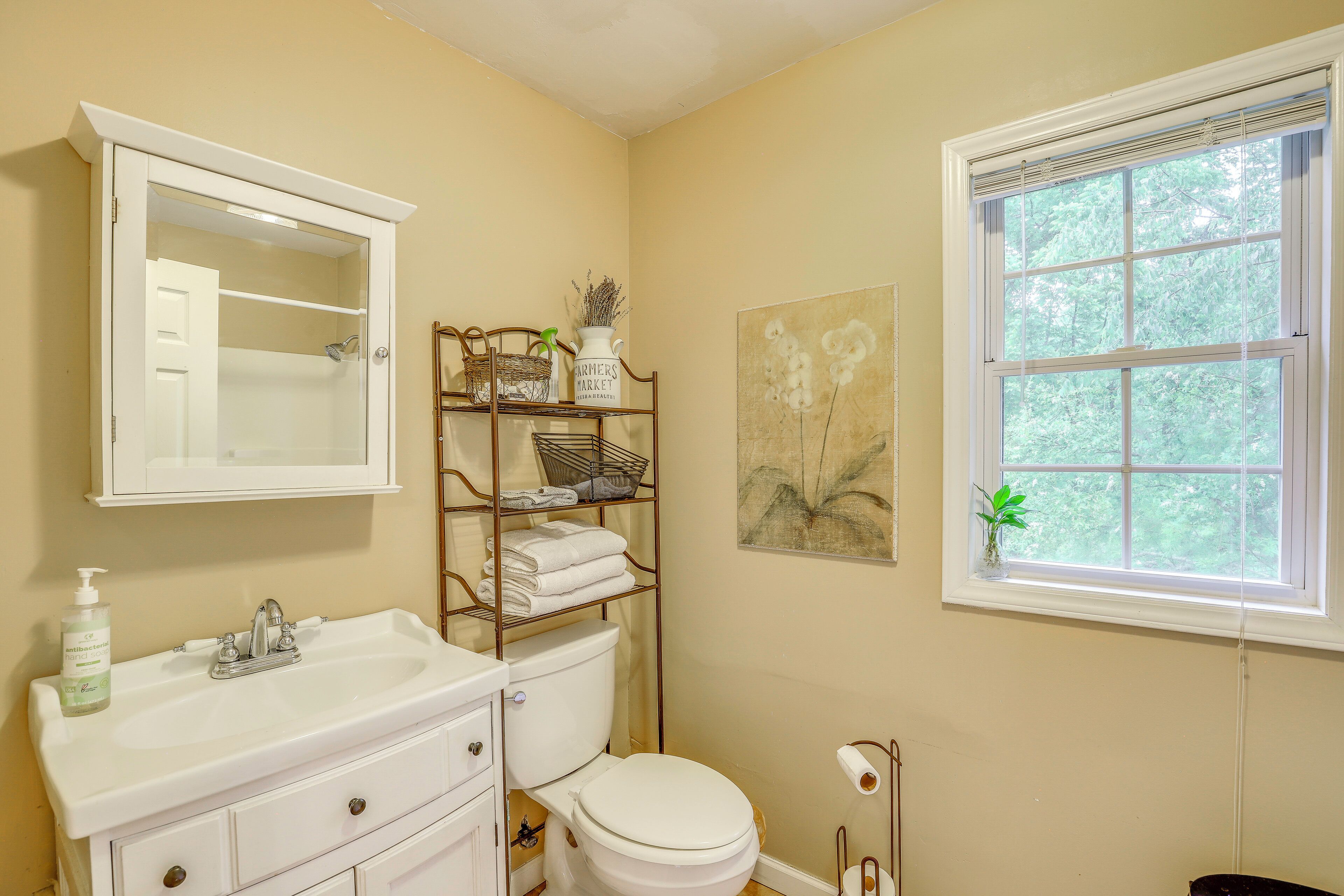 A bathroom with a white sink, a toilet, a mirror, a window with a view of trees, and a shelf with towels and a basket.
