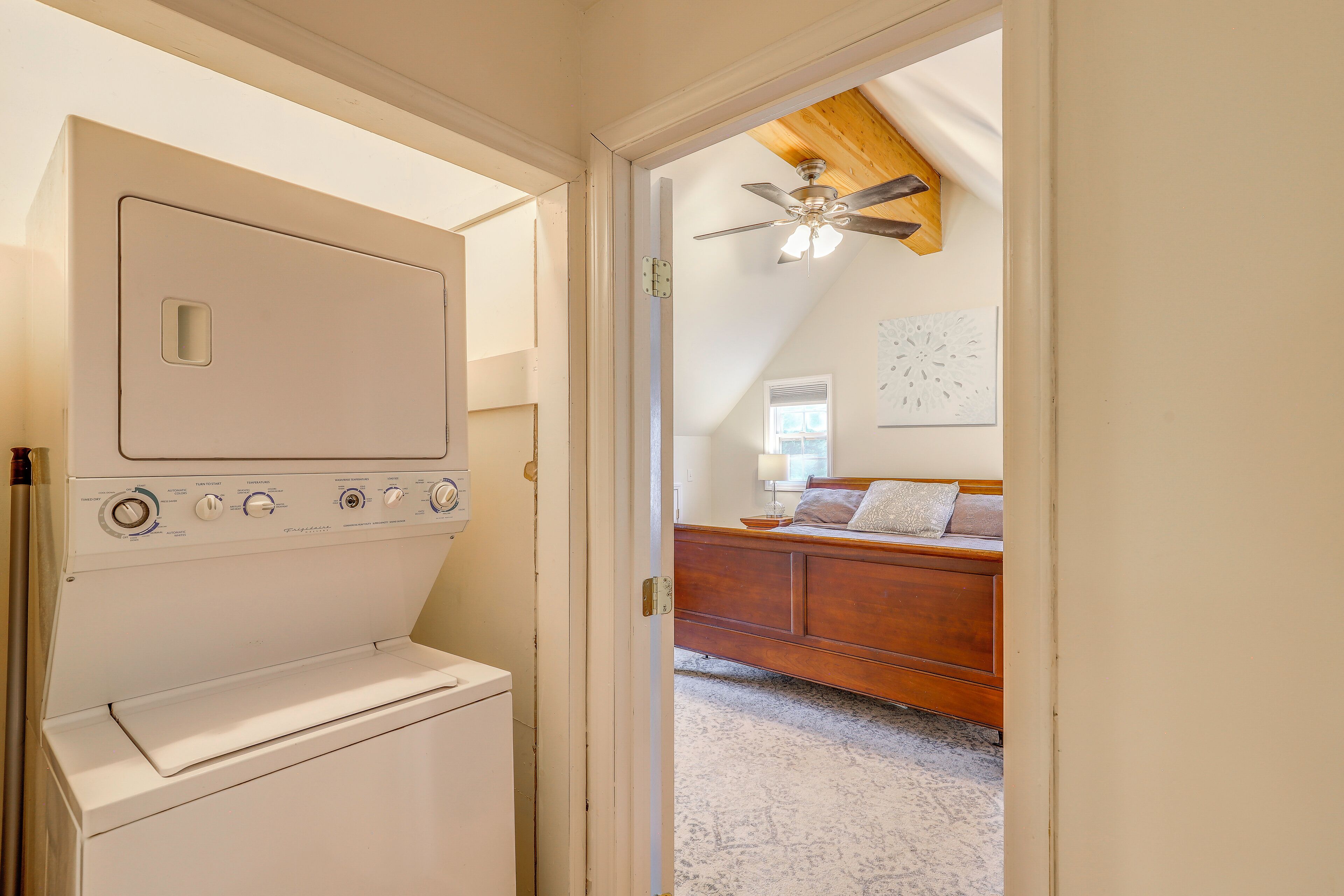 A laundry room with a washing machine, a ceiling fan, and a bedroom with a bed and a window.