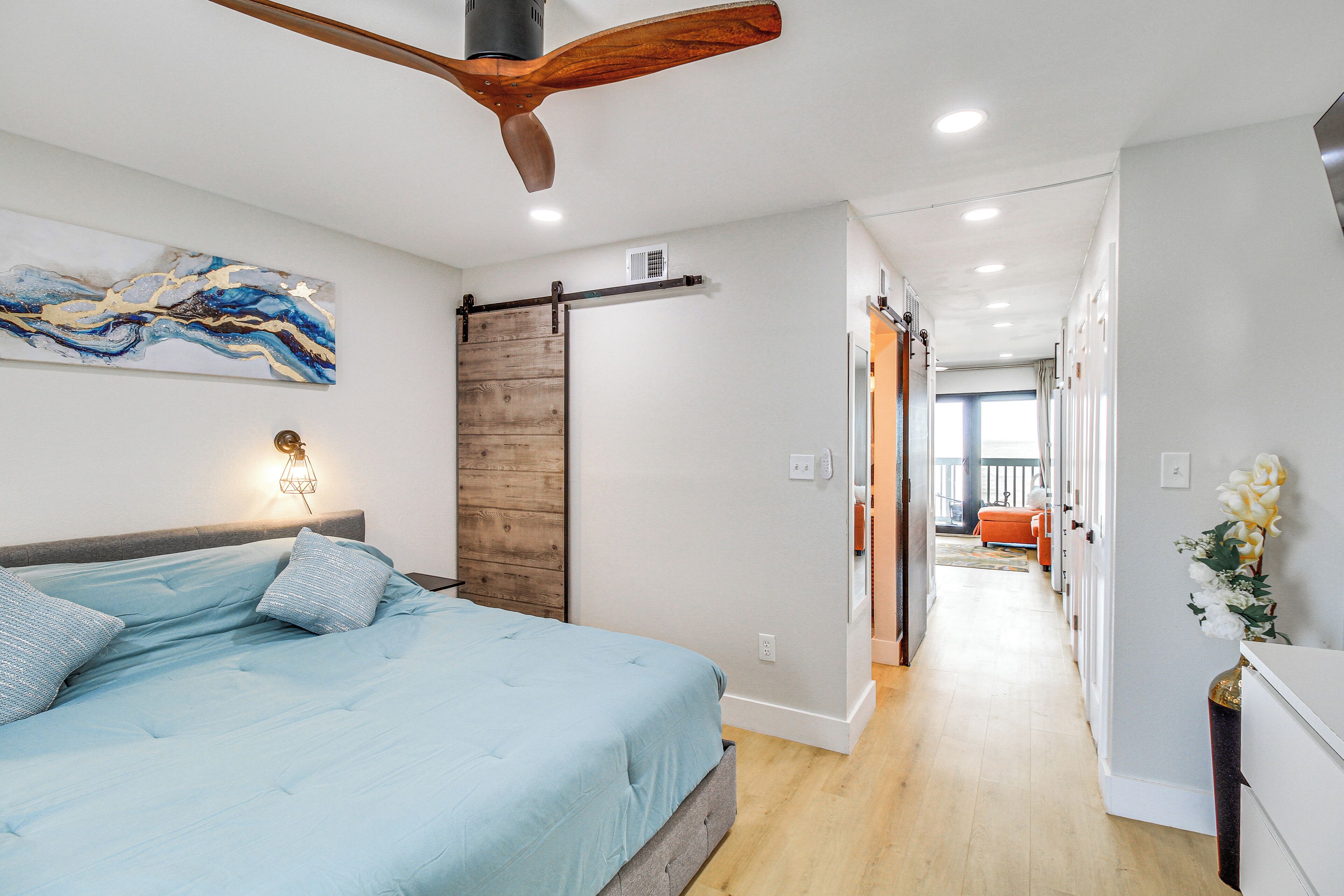 A bedroom with a bed, a wooden ceiling fan, a sliding barn door, and a hallway leading to another room.