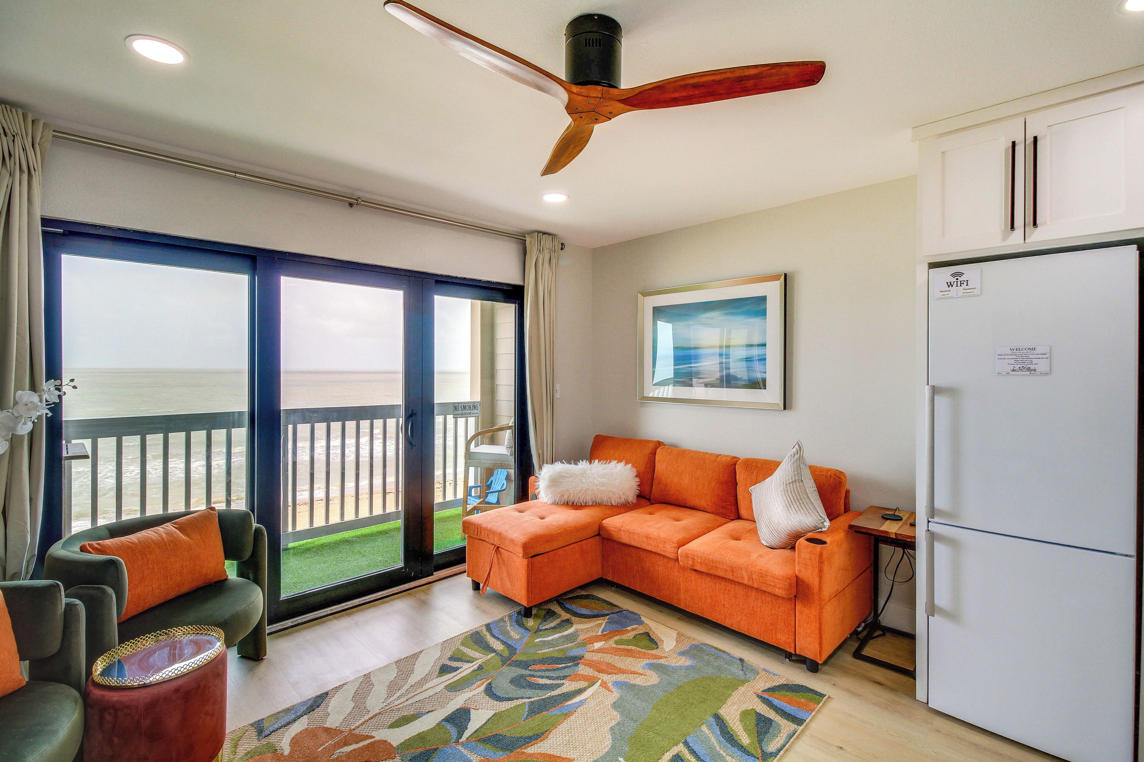 A room with a balcony, a ceiling fan, a sofa, a dining table, and a refrigerator.
