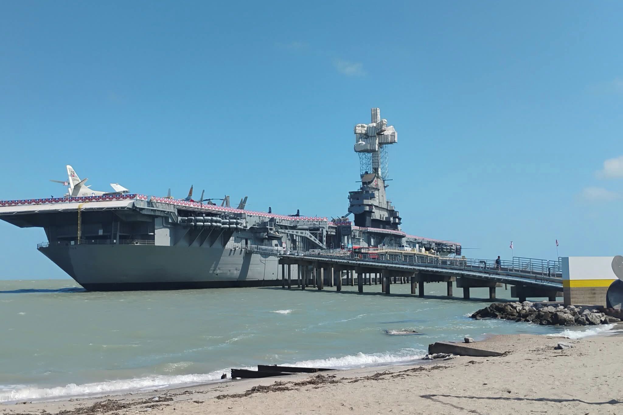 Een vliegdekschip afgemeerd aan een steiger, met een strand op de voorgrond.