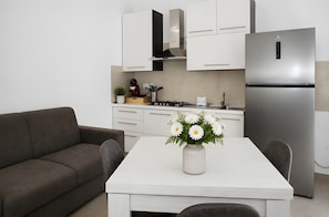 Une cuisine moderne avec des meubles blancs, un réfrigérateur en acier inoxydable, une table à manger agrémentée d’un vase de fleurs et un canapé gris.
