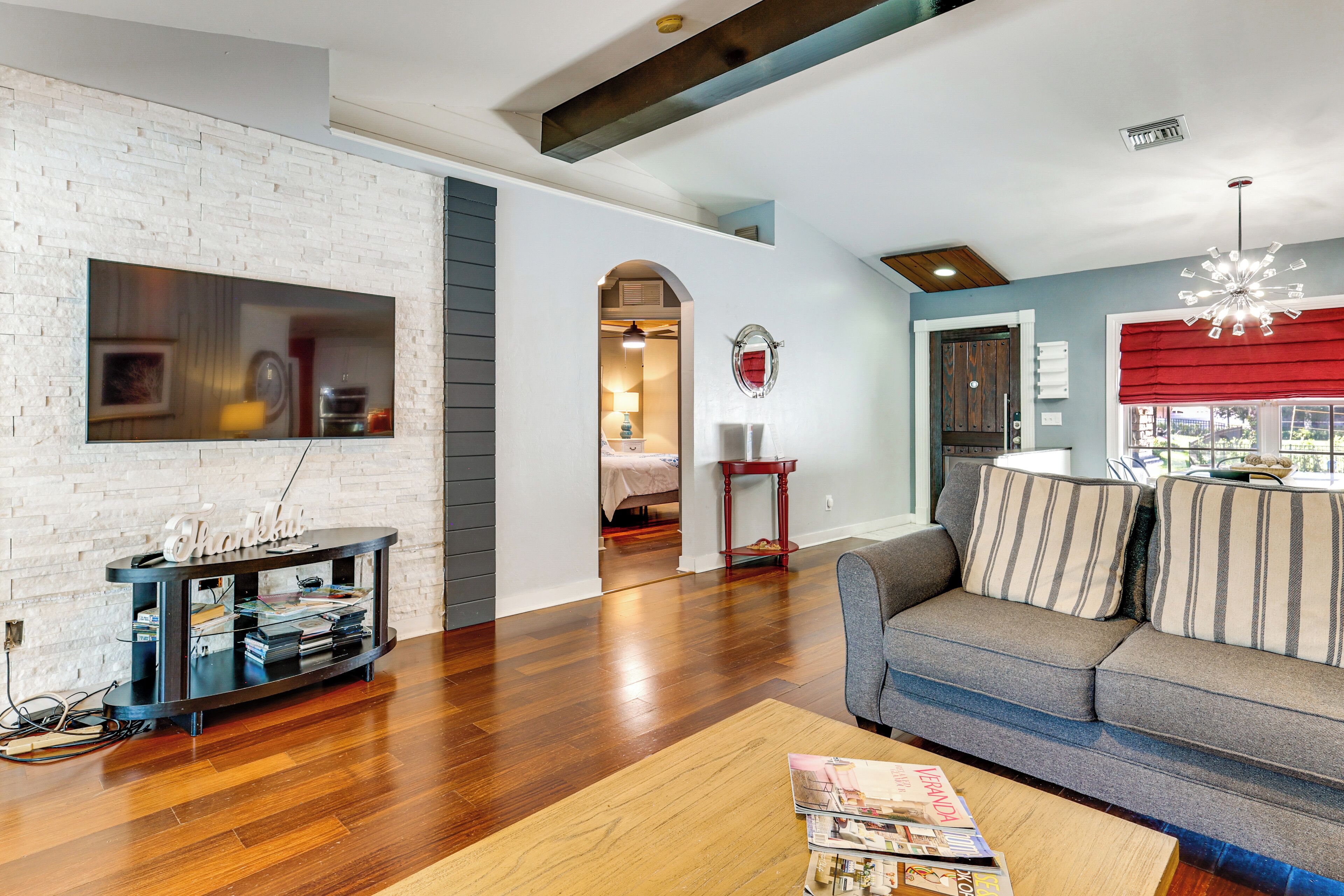 A modern living room with a flat-screen TV, a dark entertainment unit, a wooden coffee table, a gray sofa with striped cushions, and a red window valance.