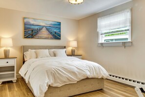 Ein Schlafzimmer mit einem Bett, zwei Nachttischen mit Lampen, einem Gemälde eines Piers und einem Fenster mit Jalousien.