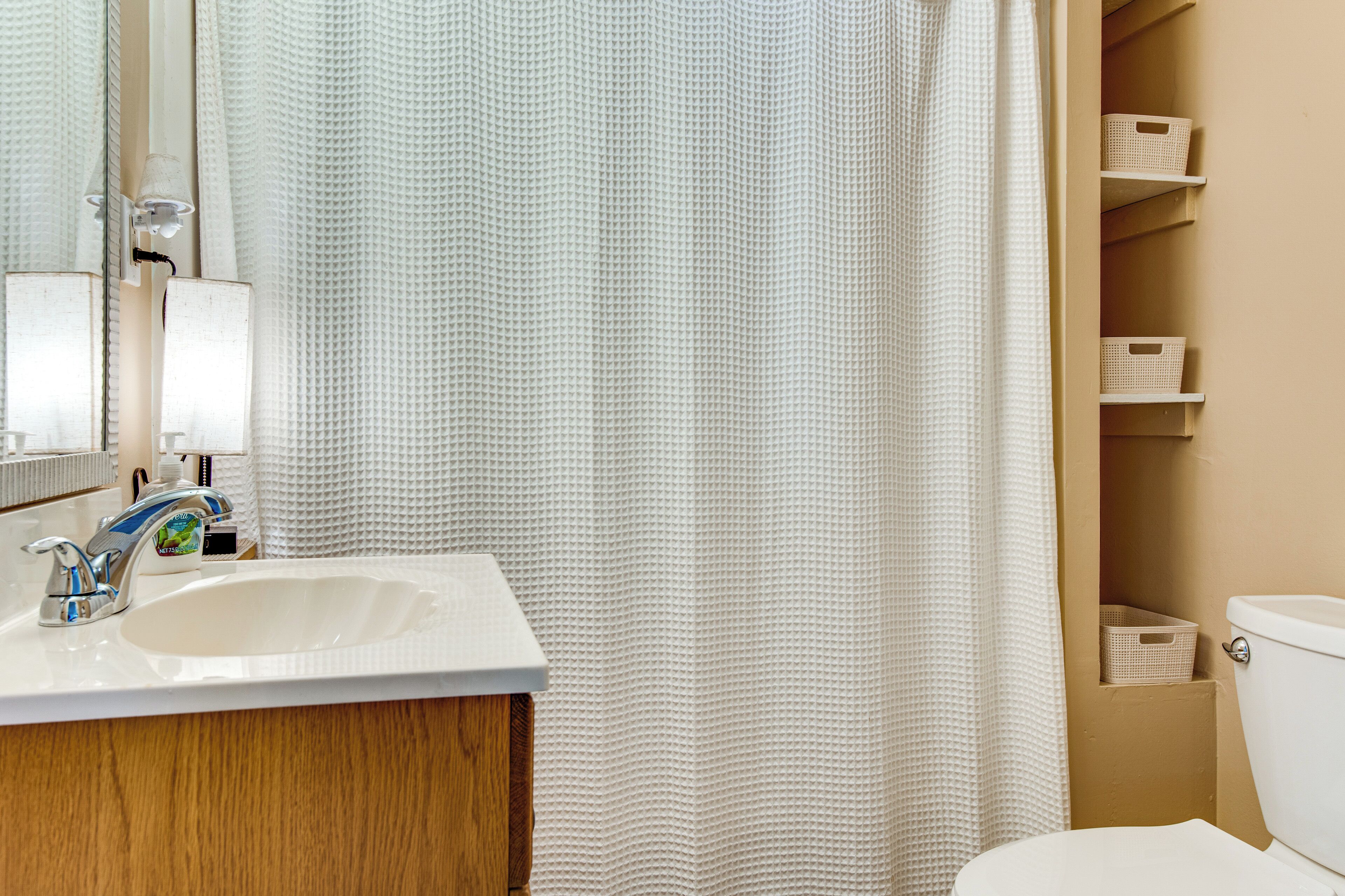 Une salle de bain avec un lavabo blanc, un miroir et un rideau de douche. Des étagères de rangement se trouvent au-dessus des toilettes.
