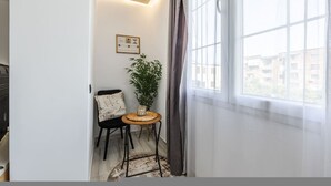 Ein Eckzimmer mit Fenster, einem kleinen Tisch mit Pflanze, einem Stuhl und einem an der Wand hängenden Bild.