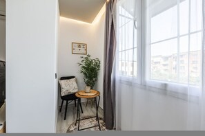 Ein Eckzimmer mit Fenster, einem kleinen Tisch mit Pflanze, einem Stuhl und einem an der Wand hängenden Bild.