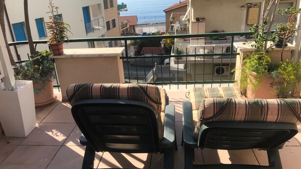 Ein Balkon mit zwei Stühlen und einem Tisch, der auf eine Küstenlandschaft mit Gebäuden und dem Meer blickt.