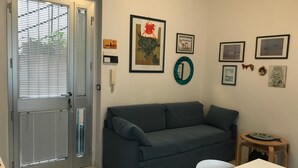 Ein Wohnzimmer mit grauem Sofa, einem weißen Tisch und einem kleinen Holzschemel. An der Wand hängen gerahmte Bilder und es gibt eine Tür mit Jalousien.