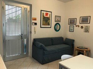 A living room with a grey sofa, a white table, and a small wooden stool. There are framed pictures on the wall and a door with blinds. - La Terrasse - Matteotti 110 (Ospedaletti)