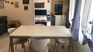 Ein kleiner, gut beleuchteter Raum mit einem weißen Tisch, zwei Stühlen, einem Bett mit gemustertem Überwurf und einem Schreibtisch mit einem Computer.